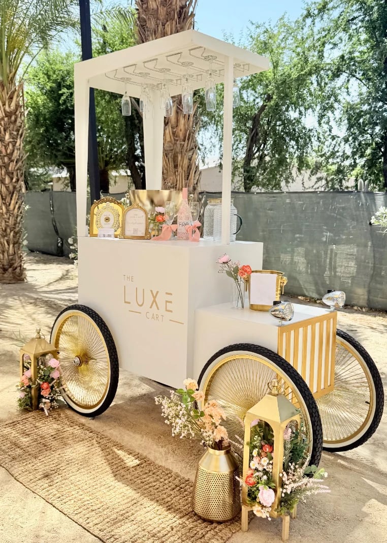 Luxe Cart vintage-style cream and gold mobile retail cart decorated with flowers and merchandise, parked in an outdoor garden setting