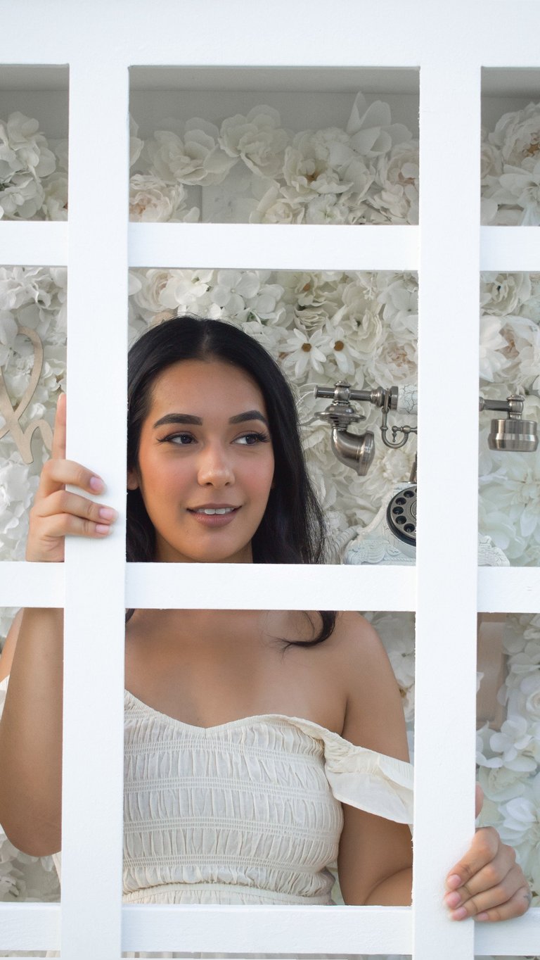 Young woman smiling at camera in white off-shoulder top with cream floral wallpaper background visible through window grid