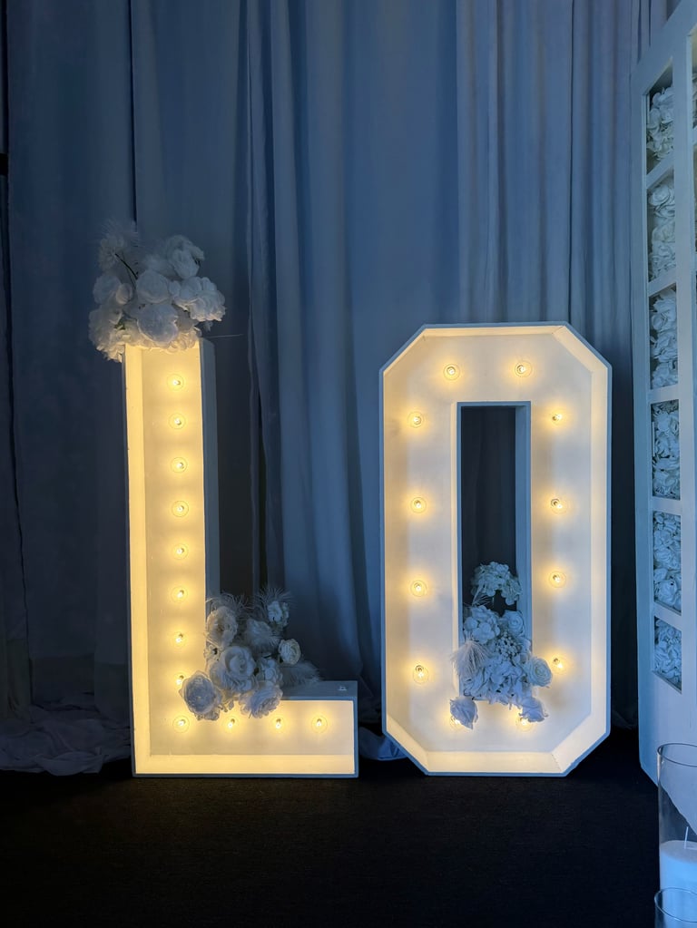 Large illuminated letters L and O with warm LED lights, decorated with white flowers against blue curtains