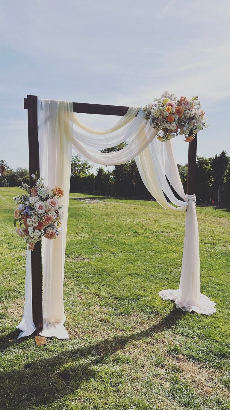 Outdoor wedding arch with white flowing fabric drapes and floral arrangements on green lawn