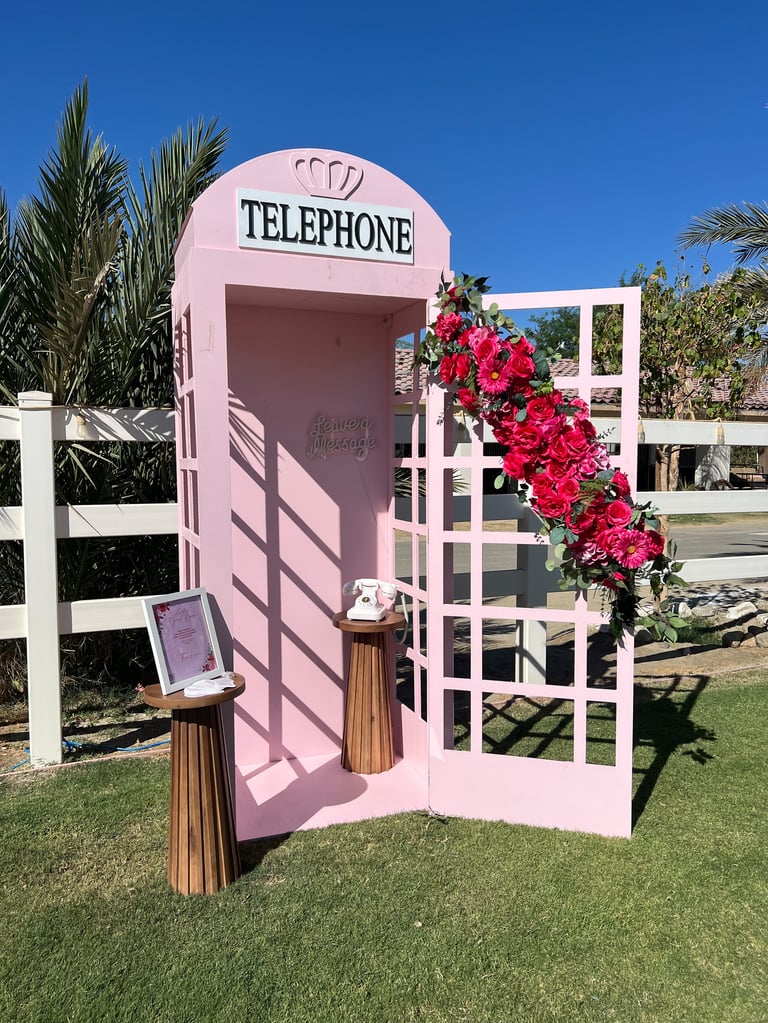 Pink vintage telephone booth with red bougainvillea flowers on lawn, palm trees and clear blue sky in background