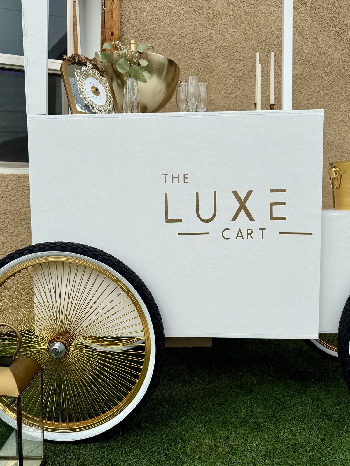 White luxury cart with gold lettering reading The Luxe Cart, featuring a large spoked wheel and decorated with gold accents and fresh flowers