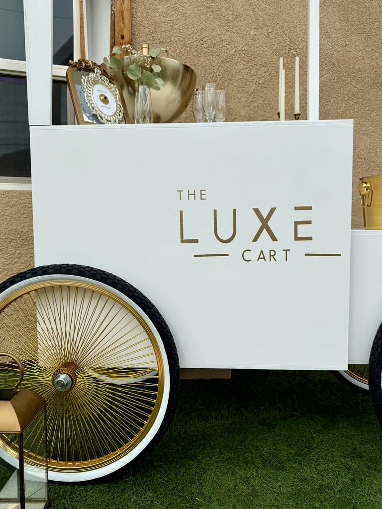 White luxury cart with gold lettering reading The Luxe Cart, featuring a large spoked wheel and decorated with gold accents and fresh flowers