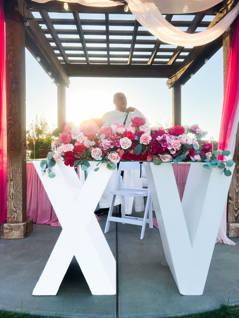 Large white X and V letters displaying vibrant pink and red floral arrangement at outdoor event under pergola with pink draping