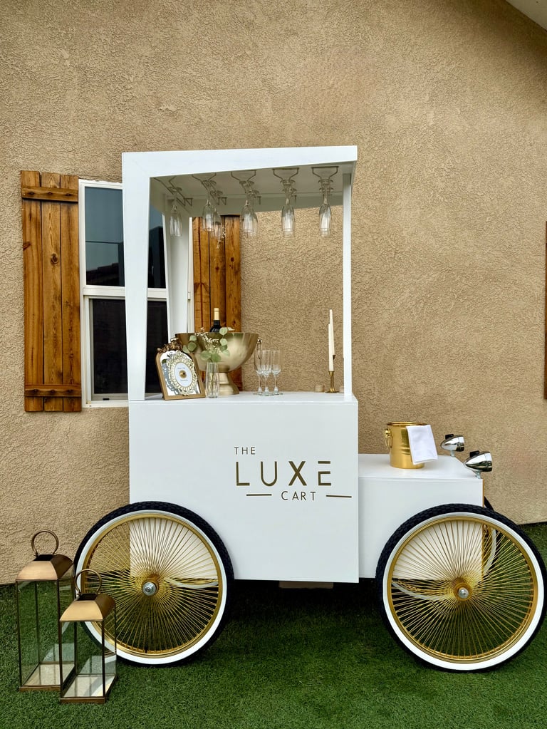 White mobile cart display labeled The Luxe Cart with large gold-trimmed wheels, serving counter, and decorative items inside.