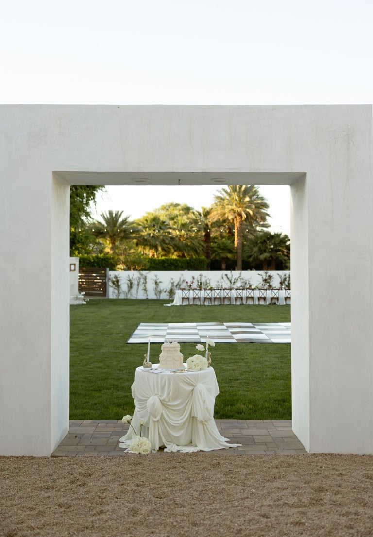 View through white archway of lawn with draped table, long seating bench, and palm trees in background