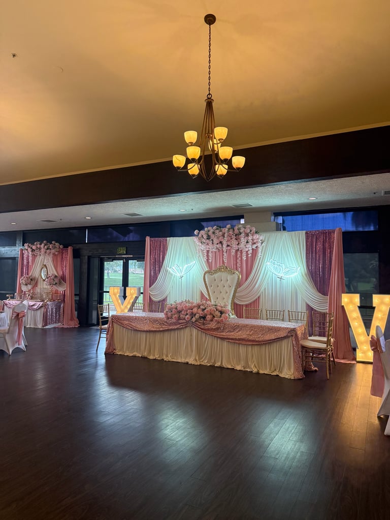 Elegant banquet hall with pink and gold wedding decorations, ornate head table, drapes, and chandelier lighting