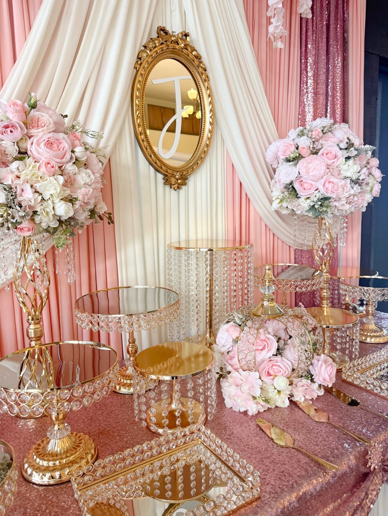 Elegant wedding display with gold ornate mirror, pink and white floral arrangements, crystal cake stands, and draped fabric backdrop.