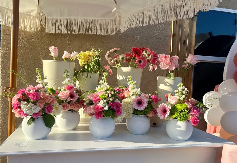 Multiple white vases with vibrant pink, red, and yellow flowers displayed on a white shelf against a gray wall