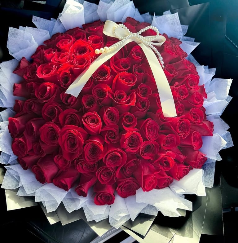 Large bouquet of deep red roses arranged in a round dome shape with white mesh wrapping and a cream-colored ribbon bow