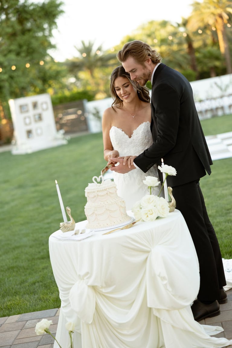 Bride and groom cutting their wedding cake at an outdoor evening reception with palm trees and garden in the background