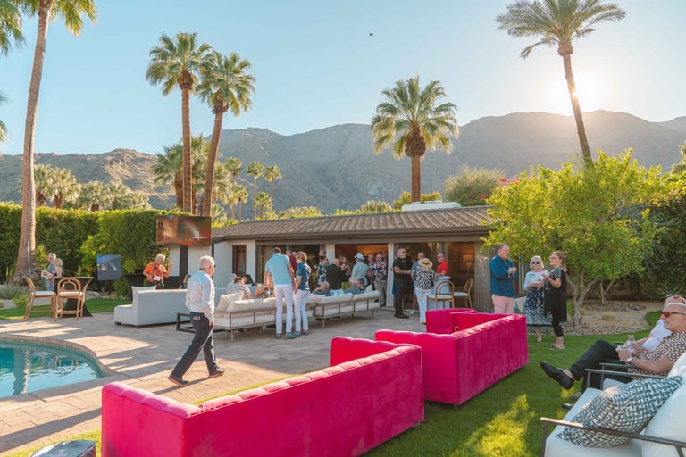Outdoor poolside event with pink seating, palm trees, mountains in background, and guests mingling near a beige pavilion.