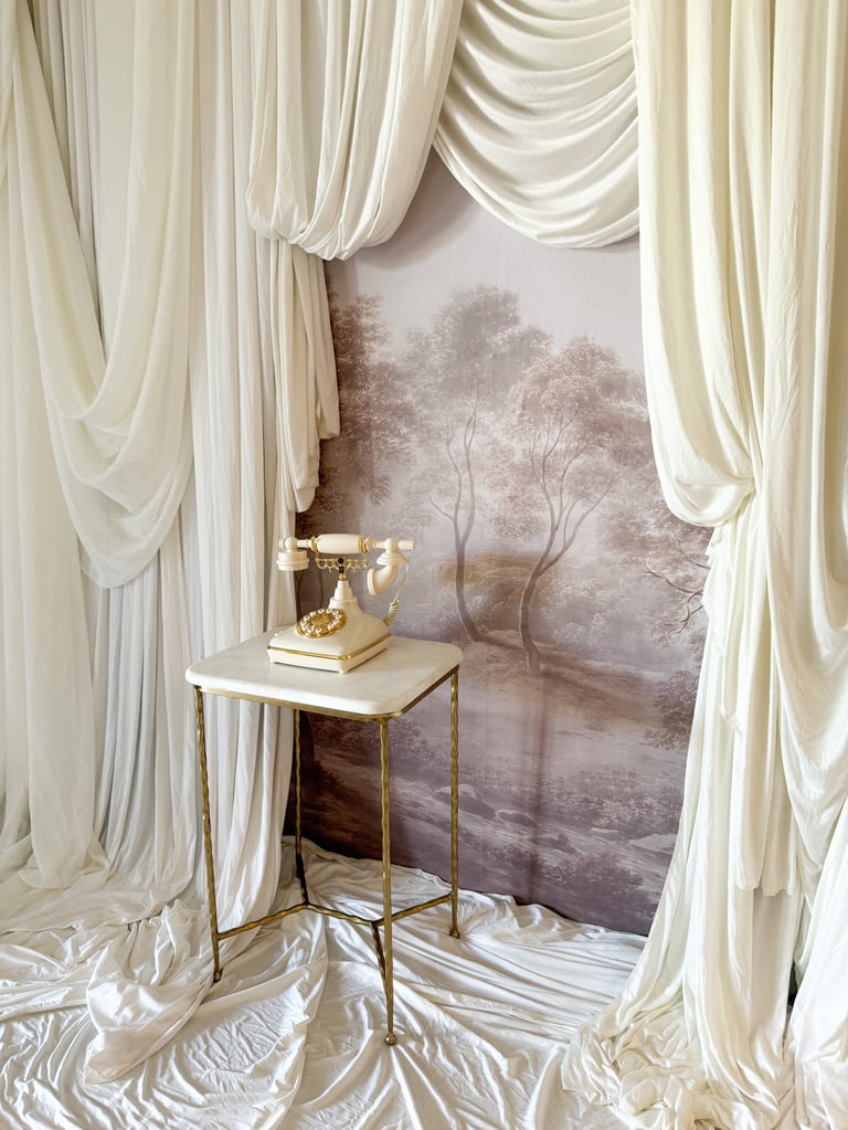Elegant cream drapes frame a marble-topped side table with a gold telephone and books near a window with misty forest scenery