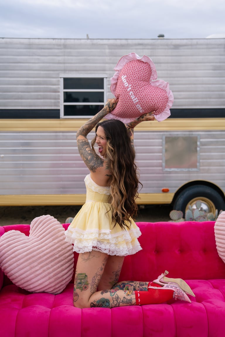 Woman with tattoos sitting on hot pink couch holding pink heart balloons in front of a vintage trailer