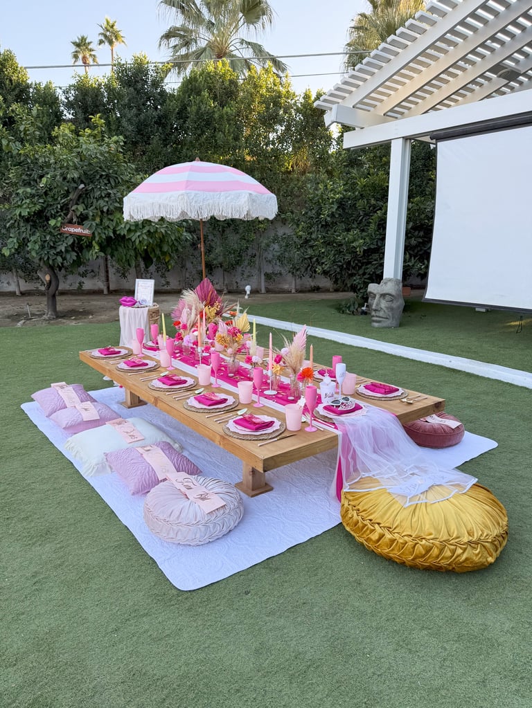 Outdoor dining setup on grass with wooden table, pink place settings, pink umbrella, round floor cushions