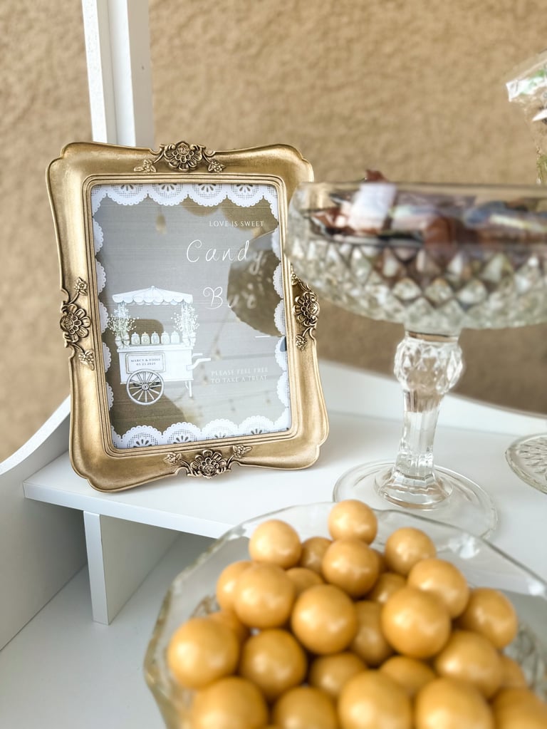 Gold ornate picture frame on white table displaying candy bar sign, with golden candies in glass bowl and decorative items in background