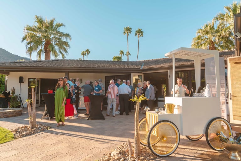 Outdoor event with guests mingling at a mid-century modern house with desert landscaping and palm trees, featuring a food cart in foreground.