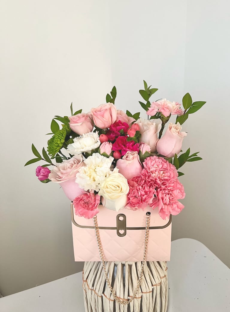 Pink floral arrangement with roses, carnations, and greenery in a pink handbag-shaped vase with chain handles on a white surface
