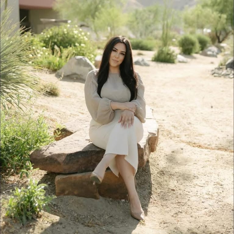 Woman with long dark hair sitting on a wooden log in a desert garden setting wearing a beige cardigan and white skirt