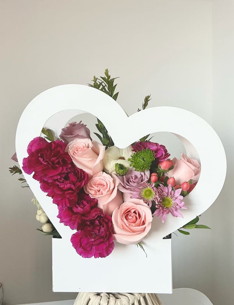 Heart-shaped white box filled with vibrant magenta carnations, pink roses, purple flowers, and green chrysanthemums with greenery