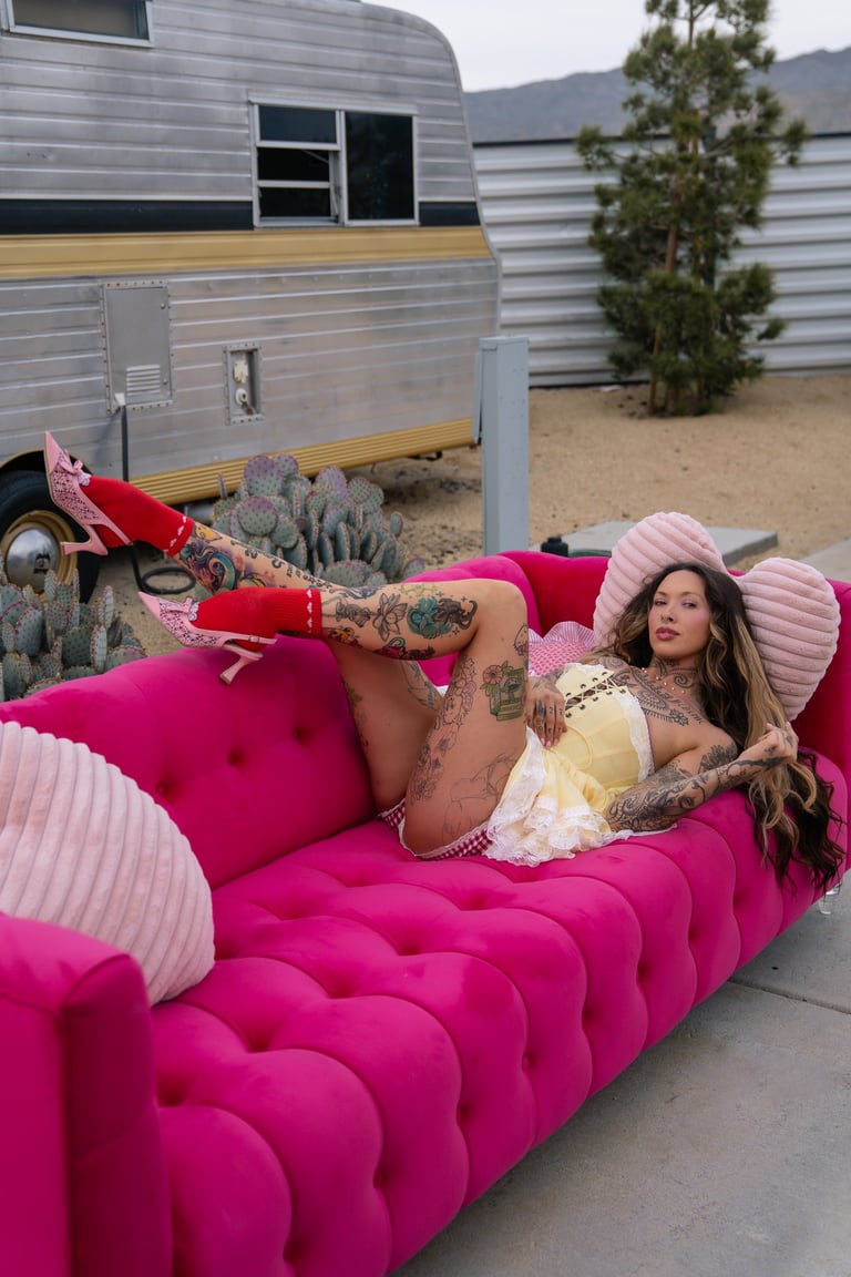 Tattooed woman reclining on a bright pink quilted bed with vintage trailers and desert plants in the background