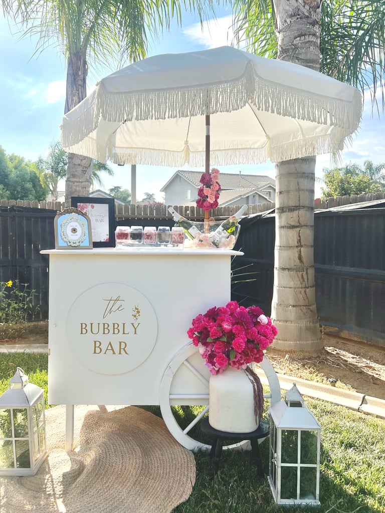 An elegant outdoor bubbly bar cart with cream-colored umbrella in a garden setting, decorated with pink flowers and surrounded by palm trees.