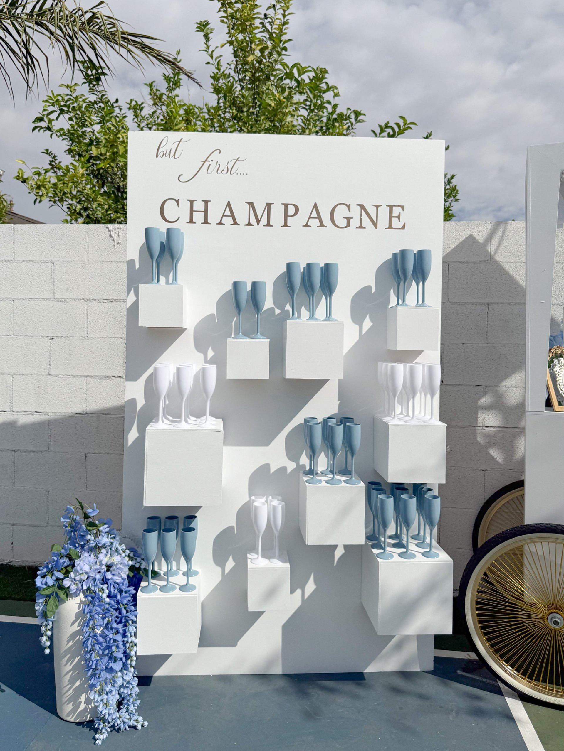 White tiered display with champagne glasses and but first Champagne text, flanked by blue flowers and decorative fan.