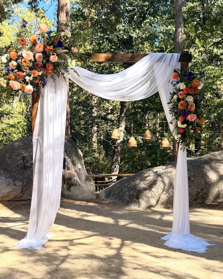 Outdoor wedding arch decorated with white draping, orange and purple flowers, and hanging lanterns in a forest setting