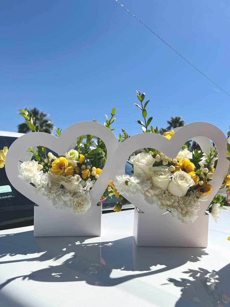 Two white heart-shaped flower arrangements with yellow and white flowers displayed outdoors in bright sunlight