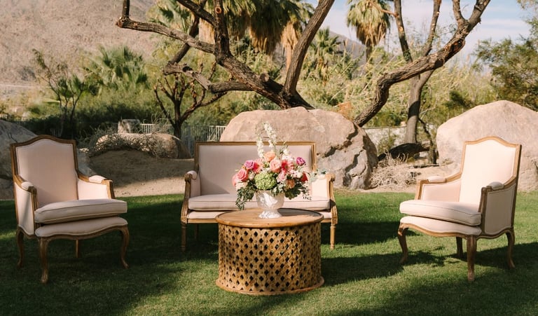 Outdoor seating arrangement with two cream armchairs facing a patterned side table with flowers, set on grass beneath large trees