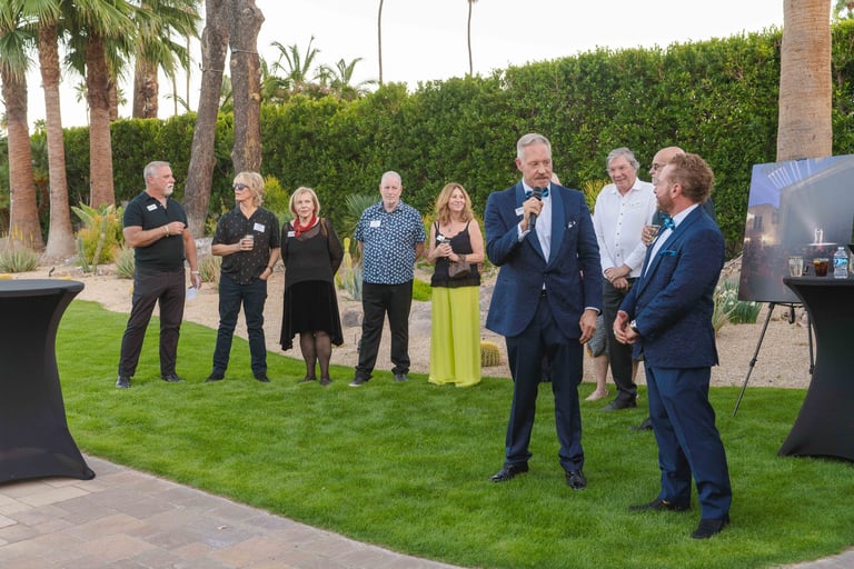Group of professionals in business attire standing on a manicured lawn at an outdoor event with palm trees and hedges in the background