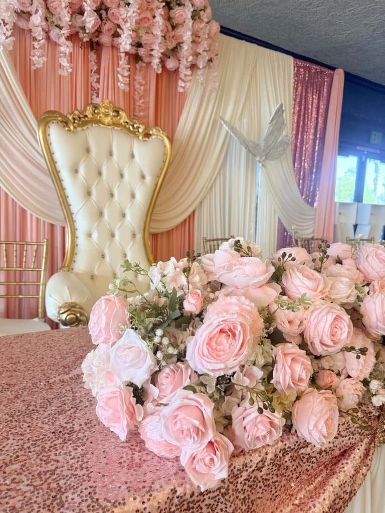 Elegant wedding reception backdrop with pink and cream drapery, hanging floral arrangements, ornate gold throne chair, and pink rose centerpiece on sequined table