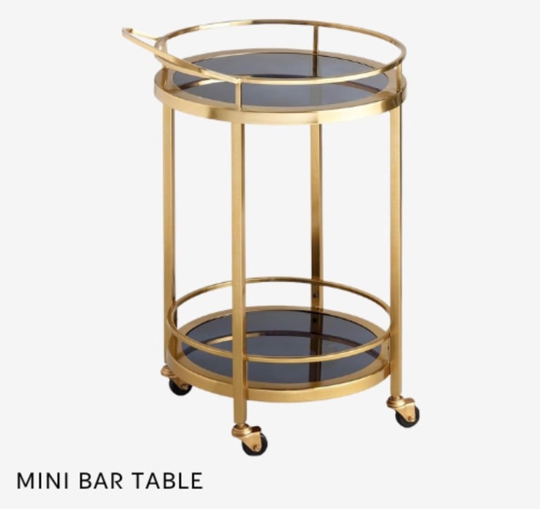 Mini bar table with brass frame, dark glass shelves, and wheeled base on white background