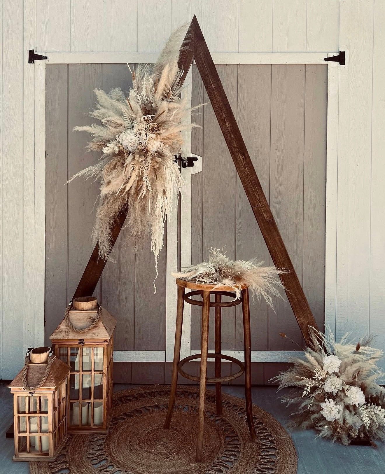 Minimalist interior display with wooden frame triangle, dried pampas grass arrangements, wooden stool, lanterns, and woven rug on neutral background