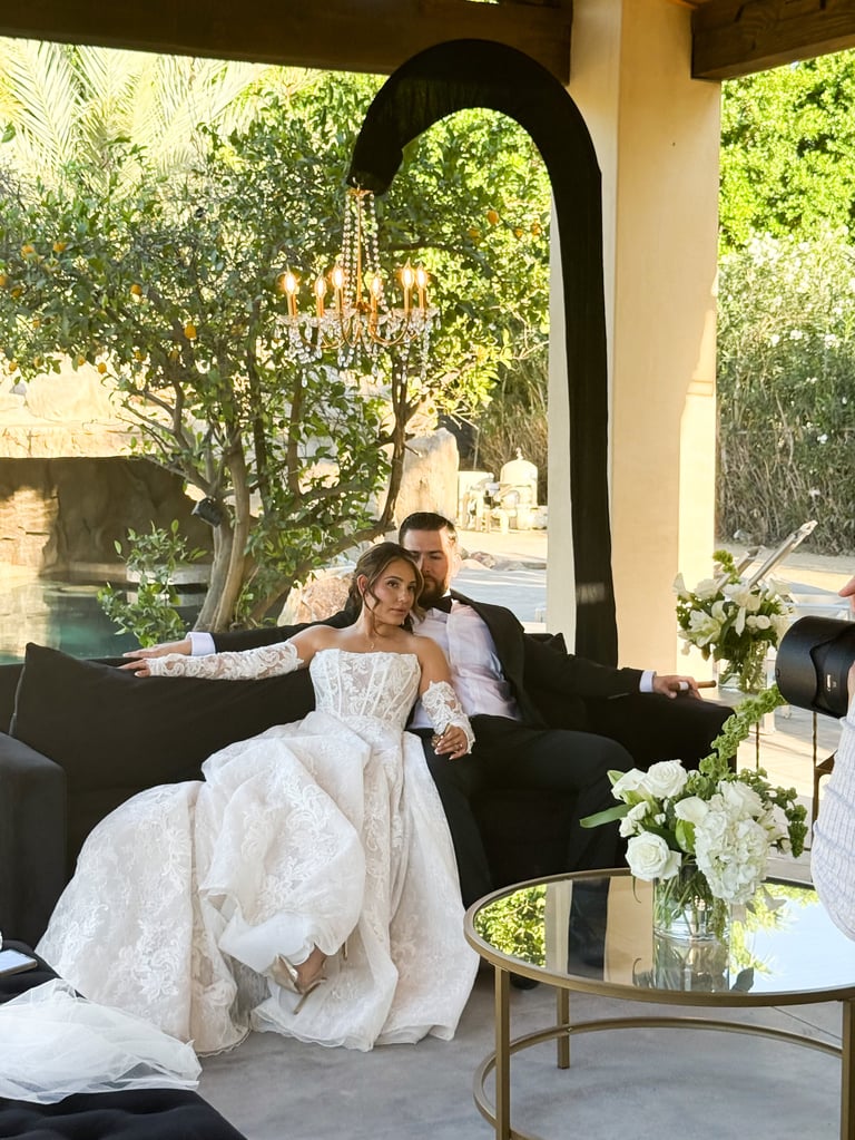 Bride and groom sitting on black sofa under arched doorway with lush green foliage and chandelier in background