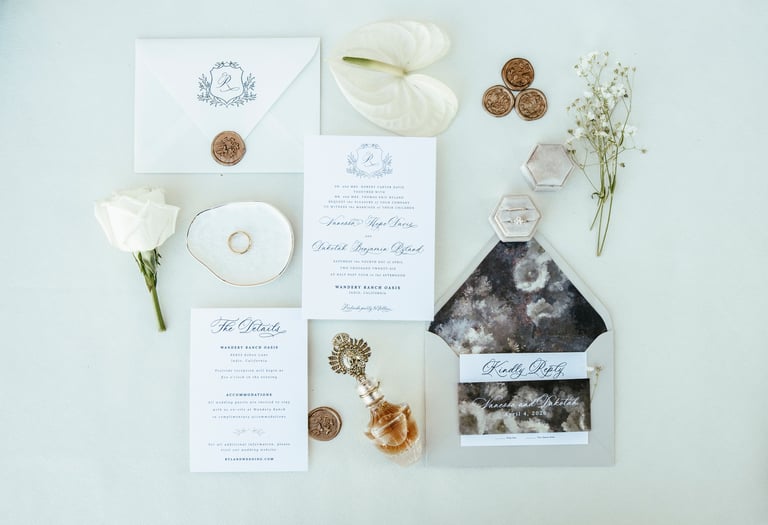 Flat lay wedding invitation suite with white stationery, gold accents, dried flowers, and wedding rings on light background