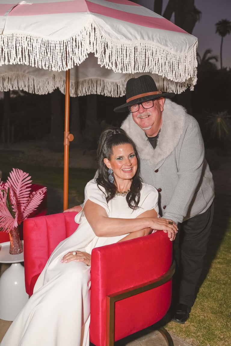 Man and woman posing together under a pink and white striped umbrella with white fringe at an outdoor evening event