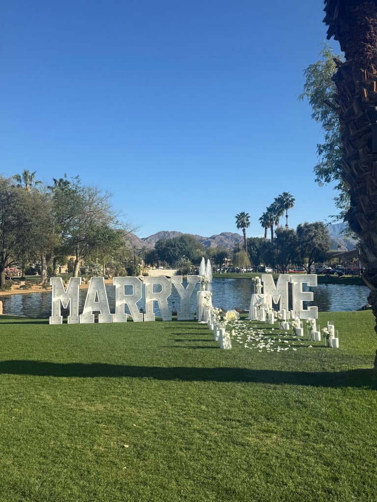 Large white MARRY ME letters on green lawn near lake with mountains and palm trees in background