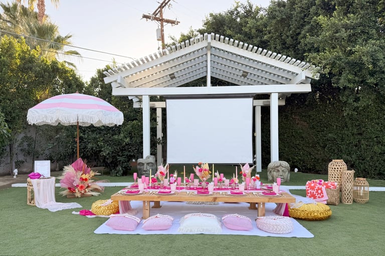 Outdoor garden event setup with pink and lavender décor, wooden table, decorative flowers and patterned cushions on lawn under white pergola and umbrella