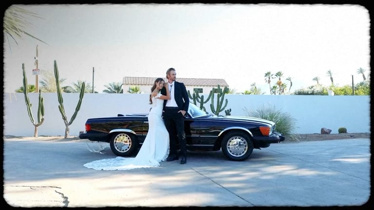 Bride and groom posing together against a classic black convertible car in front of a white wall with cacti