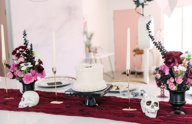 Elegant wedding or event table setting with white cake, burgundy and pink floral arrangements, candles, and decorative skull elements on burgundy runner
