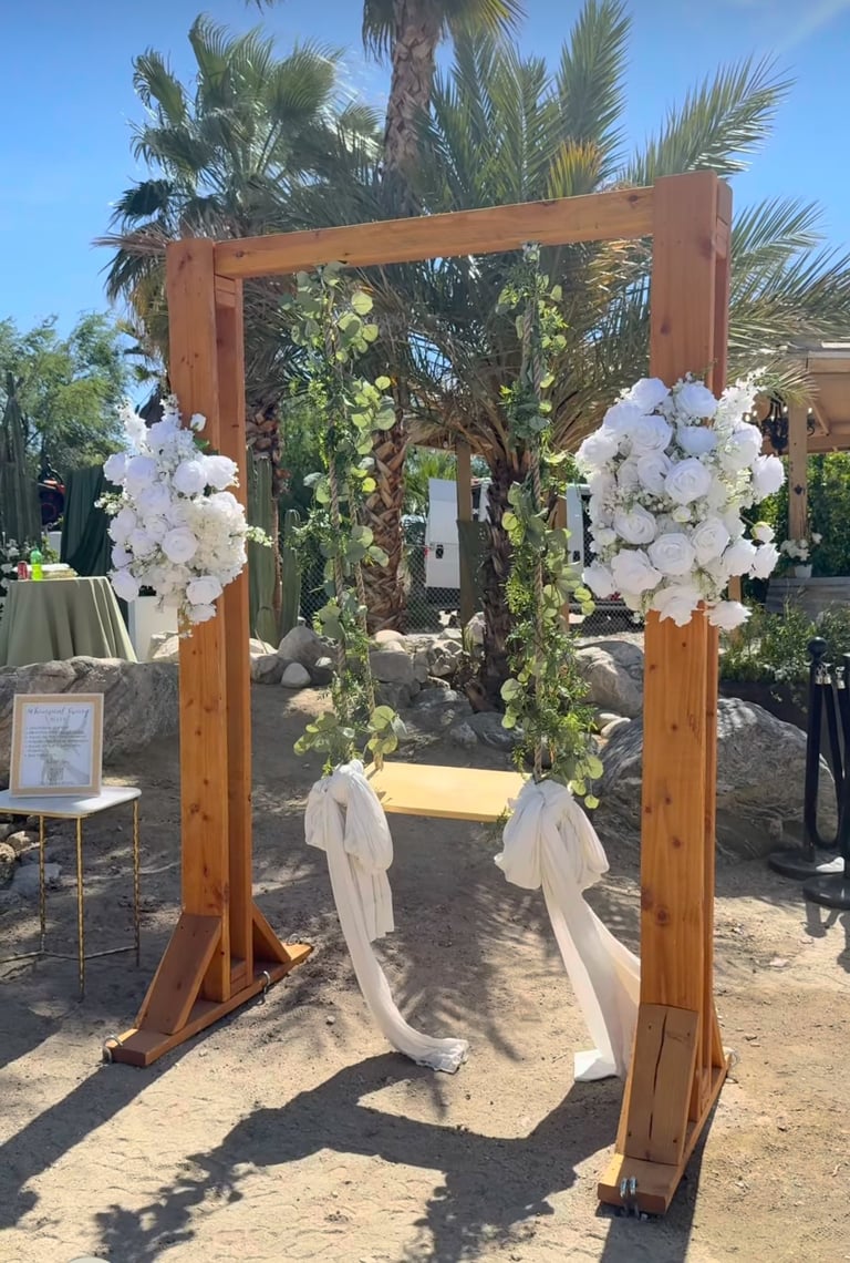 Wooden wedding arch decorated with white flowers and greenery, with palm trees in background