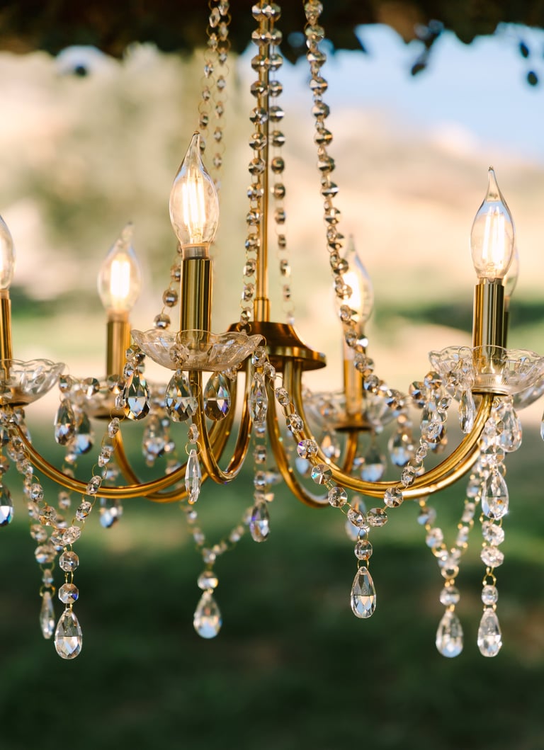 Crystal chandelier with gold accents and lit candle bulbs hanging outdoors against a blurred natural background