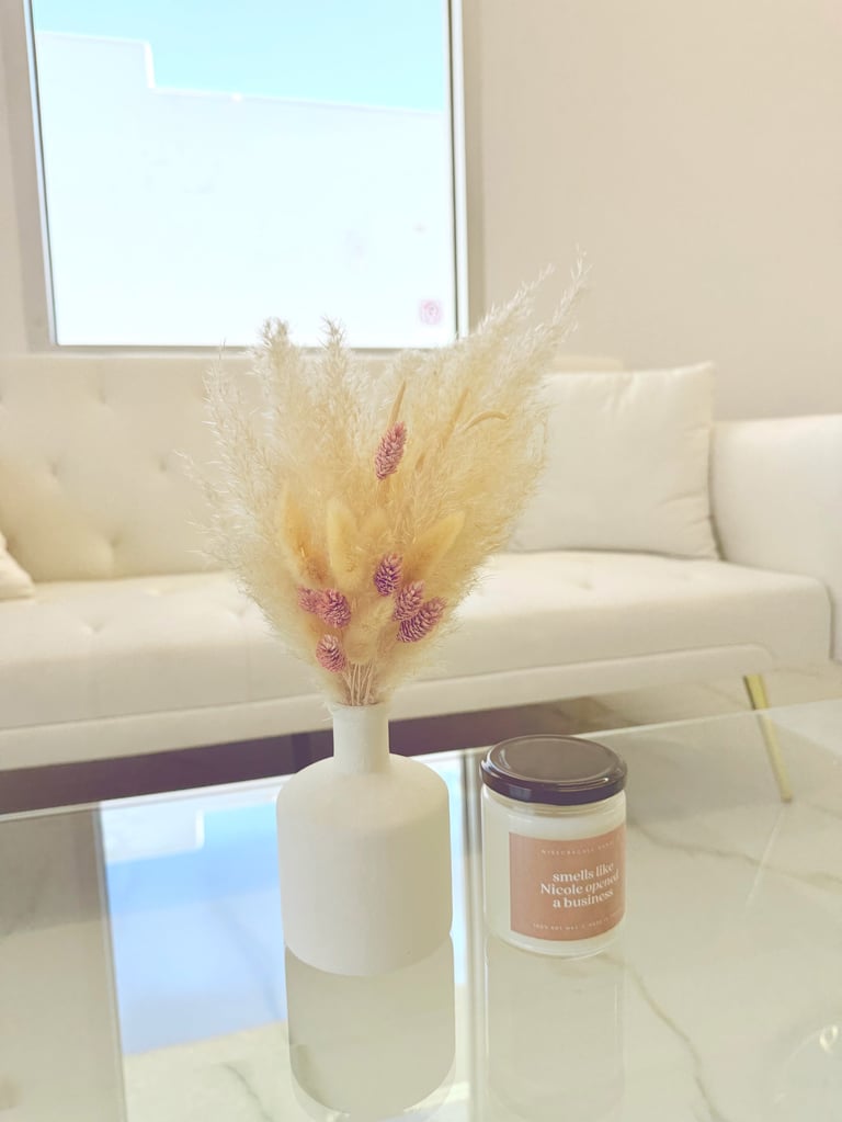 White vase with dried pampas grass and pink flowers on a glass table, next to a scented candle in a modern living room with a cream sofa.
