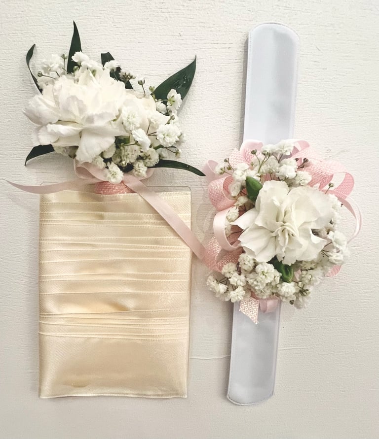 Two floral wedding corsages: one on a cream ribbon-wrapped box with white and green flowers, the other on a white rolled program with white and pink flowers.