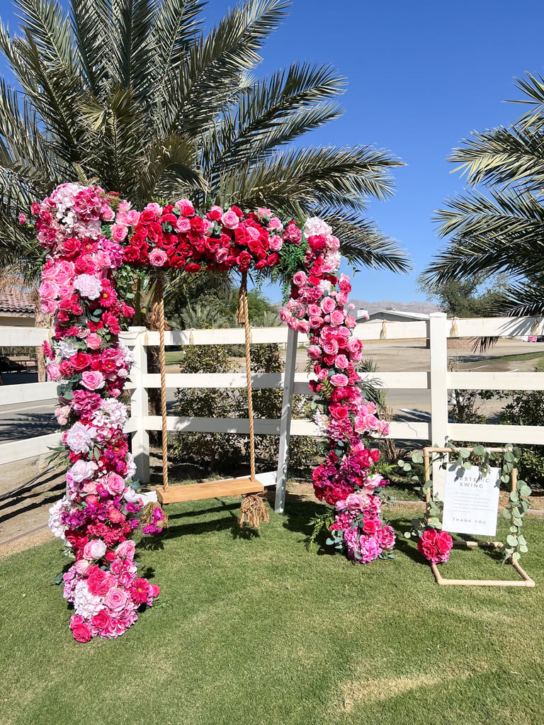 Pink and red flower archway with white wooden frame on grass, palm trees and blue sky in background