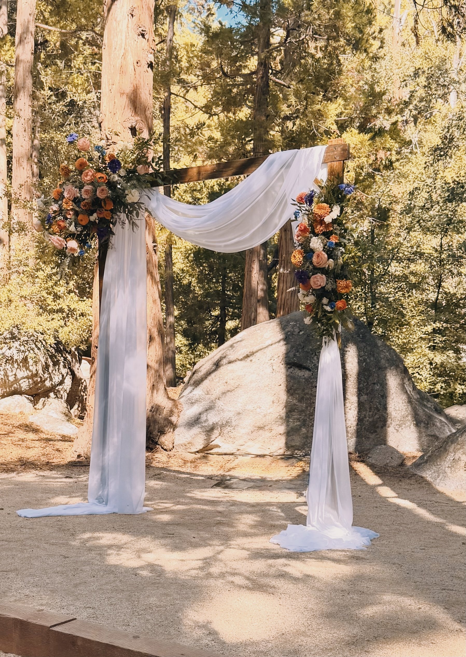 Wedding arch with white draping and orange flower arrangements in a forest setting