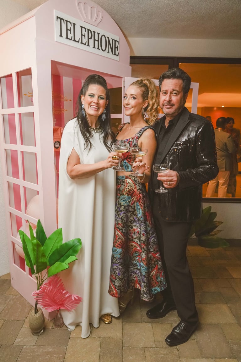 Three people posing with drinks next to a pink British telephone booth at an indoor event