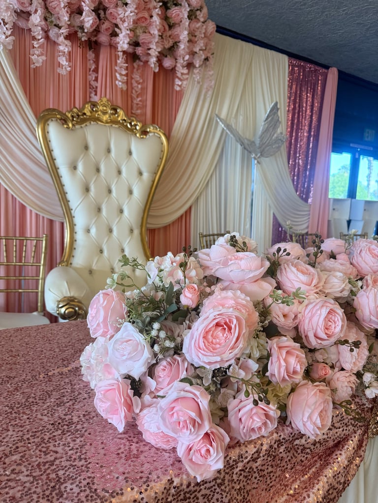 Elegant wedding reception setup with pink roses, ornate gold-framed chair, and flowing pink and white draped fabric backdrop