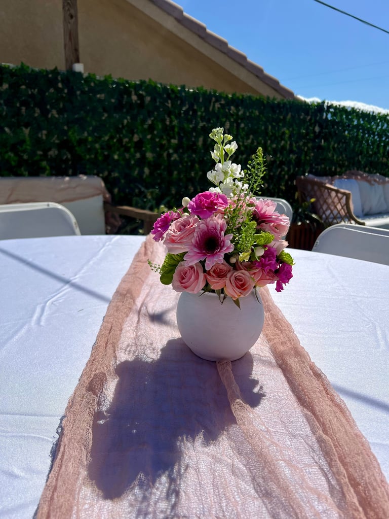 Floral arrangement with pink and magenta flowers in white vase on outdoor patio table with green hedge in background